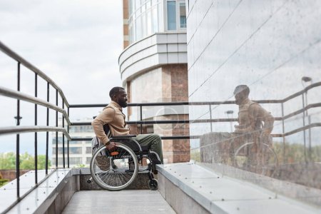 Foto de Vista lateral de longitud completa del hombre con discapacidad empujando rueda de la silla de ruedas mientras que va por la rampa en la ciudad, espacio de copia - Imagen libre de derechos