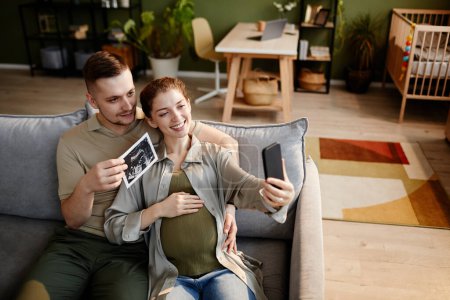 Foto de Feliz joven pareja hablando sobre el embarazo en línea con sus familiares utilizando el teléfono móvil mientras está sentado en el sofá en casa - Imagen libre de derechos