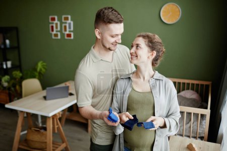 Téléchargez les photos : Jeune homme aidant sa femme enceinte à mettre des choses pour enfants dans la chambre, ils se préparent à la naissance de l'enfant - en image libre de droit
