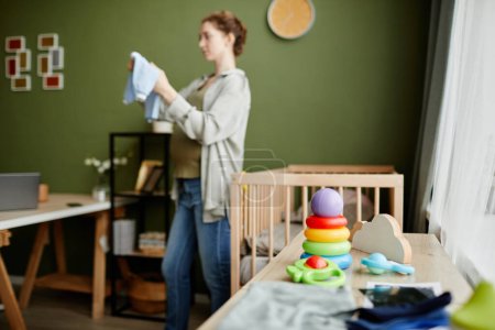 Téléchargez les photos : Image horizontale de vêtements et jouets pour nouveau-né sur commode avec femme enceinte préparant des choses pour l'accouchement en arrière-plan - en image libre de droit