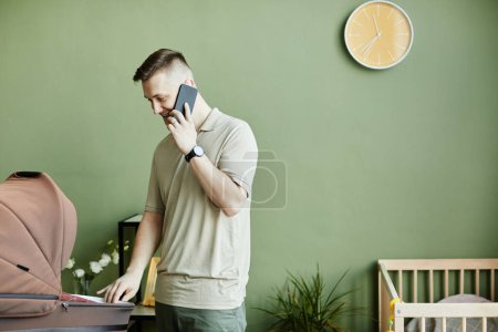 Photo for Young dad talking on mobile phone while babysitting with his baby in pram in the room - Royalty Free Image