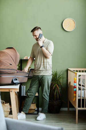 Foto de Hombre joven hablando por teléfono móvil mientras se preocupa por su bebé recién nacido en la cuna en casa - Imagen libre de derechos