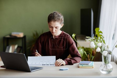 Foto de Joven empresaria sentada a la mesa con portátil y utilizando el calendario para planificar su trabajo en casa - Imagen libre de derechos