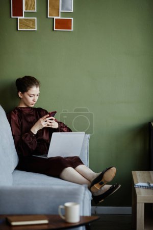 Téléchargez les photos : Jeune femme assise sur un canapé avec un ordinateur portable sur les genoux et utilisant le téléphone mobile pour le travail en ligne à la maison - en image libre de droit
