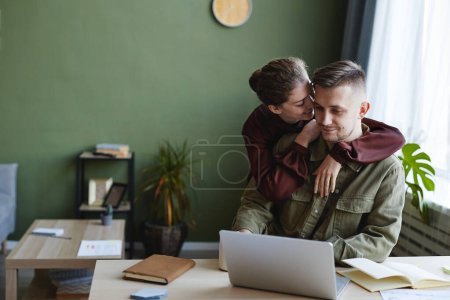 Téléchargez les photos : Jeune femme embrassant son homme derrière pendant qu'il travaille sur ordinateur portable à la table à la maison - en image libre de droit
