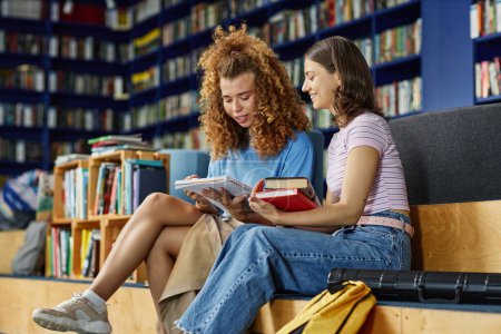 Foto de Vibrante retrato de dos chicas jóvenes leyendo libros en la biblioteca de la universidad y sonriendo, espacio para copiar - Imagen libre de derechos
