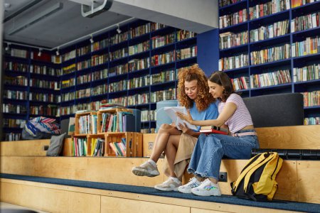 Téléchargez les photos : Vibrant portrait pleine longueur de deux adolescentes lisant des livres dans la bibliothèque de l'université et souriant, espace de copie - en image libre de droit