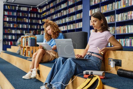 Téléchargez les photos : Vue de côté portrait d'adolescente moderne utilisant un ordinateur portable dans la bibliothèque de l'école et étudier, copier l'espace - en image libre de droit