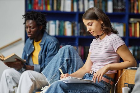 Téléchargez les photos : Vue latérale vibrante portrait de jeune femme faisant des devoirs à la bibliothèque du collège et écrivant dans un cahier - en image libre de droit