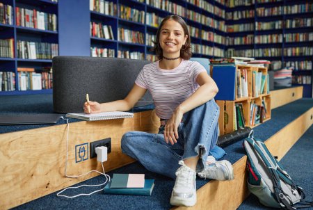 Téléchargez les photos : Vibrant portrait pleine longueur d'une étudiante souriante assise les jambes croisées dans la bibliothèque de l'école, espace de copie - en image libre de droit