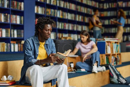 Téléchargez les photos : Portrait de jeune homme noir lisant un livre dans le salon de la bibliothèque avec des étudiants en arrière-plan, espace de copie - en image libre de droit