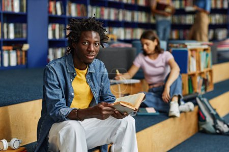 Téléchargez les photos : Portrait vibrant du jeune homme noir lisant le livre dans la bibliothèque du collège et souriant à la caméra, espace de copie - en image libre de droit