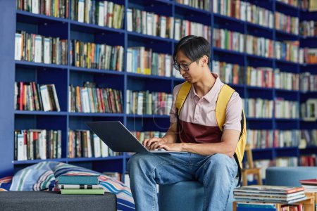 Téléchargez les photos : Portrait de jeune homme asiatique utilisant un ordinateur portable dans le salon de la bibliothèque avec des tons bleus vibrants, espace de copie - en image libre de droit