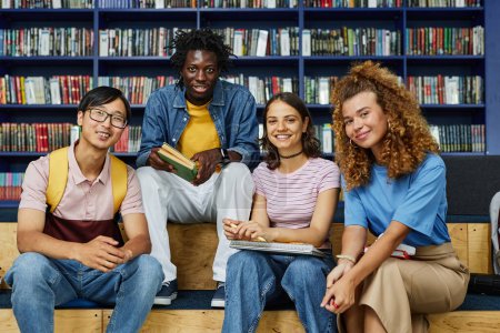 Foto de Vista frontal al diverso grupo de estudiantes en la biblioteca sonriendo felizmente a la cámara contra las estanterías - Imagen libre de derechos