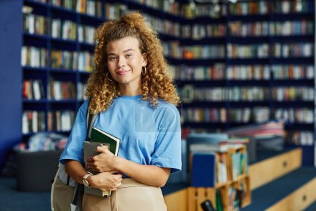 Téléchargez les photos : Taille vibrante portrait de cheveux bouclés jeune femme tenant des livres dans la bibliothèque de l'université et regardant la caméra, espace de copie - en image libre de droit