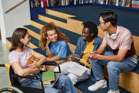 Téléchargez les photos : Groupe diversifié de jeunes discutant du travail à la bibliothèque du collège, y compris les étudiantes handicapées - en image libre de droit
