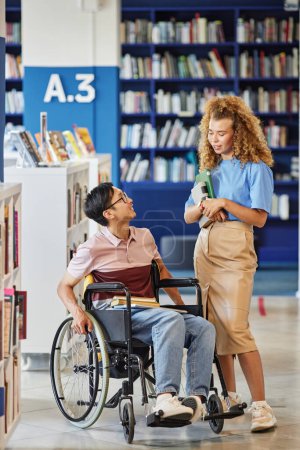 Téléchargez les photos : Portrait vertical complet d'un étudiant asiatique handicapé parlant à un ami dans un cadre de bibliothèque collégiale - en image libre de droit