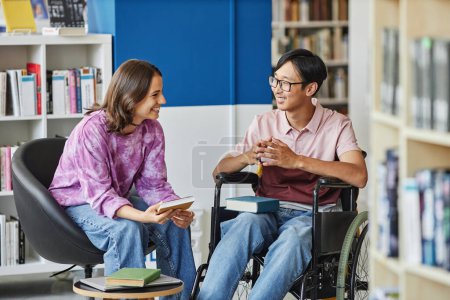Foto de Retrato de un joven sonriente en silla de ruedas hablando con un amigo mientras estudian juntos en la biblioteca - Imagen libre de derechos