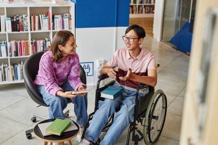 Téléchargez les photos : Portrait grand angle de jeune homme asiatique en fauteuil roulant parlant à un ami tout en étudiant ensemble dans la bibliothèque - en image libre de droit