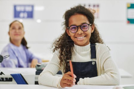 Foto de Retrato de una colegiala negra sonriente con gafas sentada en el escritorio del aula y mirando a la cámara - Imagen libre de derechos