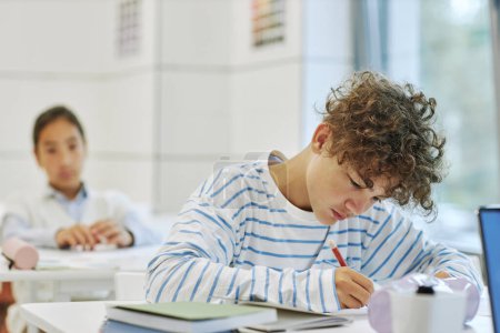 Téléchargez les photos : Portrait de jeune garçon aux cheveux bouclés assis au bureau dans la classe de l'école et écrivant, espace de copie - en image libre de droit