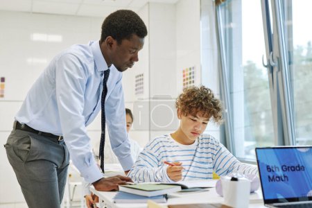 Photo for Side view portrait of young black teacher helping boy in school classroom - Royalty Free Image