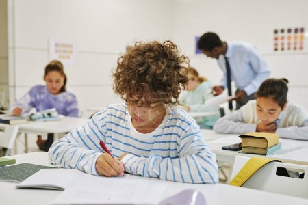 Foto de Retrato de un joven colegial con el pelo rizado sentado en el escritorio en el aula de la escuela y escribiendo en un cuaderno - Imagen libre de derechos
