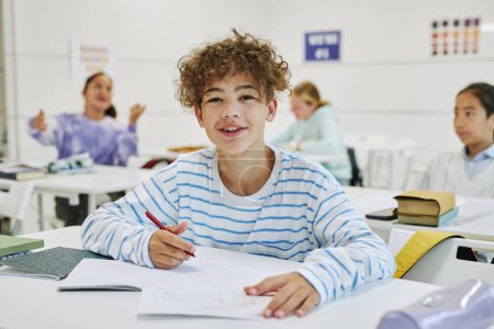 Téléchargez les photos : Portrait de jeune écolier aux cheveux bouclés assis au bureau dans la classe de l'école et souriant à la caméra - en image libre de droit