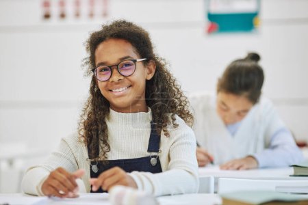 Photo for Portrait of smiling black schoolgirl wearing glasses and looking at camera while sitting at desk in school, copy space - Royalty Free Image