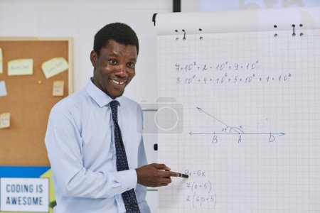 Téléchargez les photos : Portrait taille haute du jeune professeur noir expliquant problème de mathématiques à la classe et pointant vers le tableau à feuilles mobiles - en image libre de droit