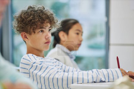 Foto de Retrato de vista lateral de adolescente multiétnico con el pelo rizado sentado en el escritorio en la escuela y escuchando a la maestra - Imagen libre de derechos
