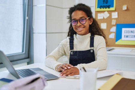Foto de Retrato de colegiala negra sonriente sentada en el escritorio con portátil en el aula de la escuela - Imagen libre de derechos