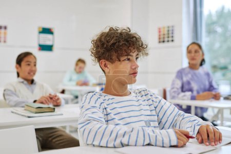 Téléchargez les photos : Portrait d'un adolescent assis au bureau dans une classe d'école et écoutant un enseignant avec un groupe d'enfants en arrière-plan - en image libre de droit