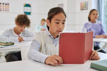 Téléchargez les photos : Portrait d'un adolescent assis au bureau dans une classe d'école et lisant un livre avec un groupe d'enfants en arrière-plan - en image libre de droit