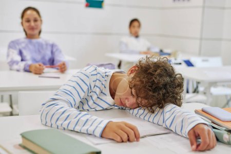 Téléchargez les photos : Portrait d'un jeune garçon dormant sur le bureau dans une salle de classe avec des enfants en arrière-plan - en image libre de droit