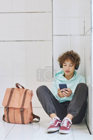 Photo for Minimal full length portrait of isolated teen schoolboy sitting alone in corner and using smartphone - Royalty Free Image