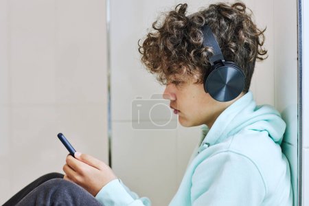 Photo for Minimal side view portrait of teen schoolboy sitting on floor and using smartphone with headphones - Royalty Free Image