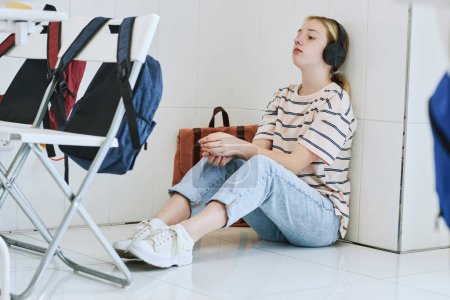 Téléchargez les photos : Portrait pleine longueur d'adolescente déprimée assise sur le sol à l'école avec écouteurs - en image libre de droit