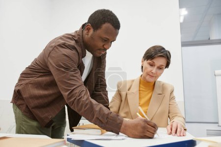 Téléchargez les photos : Femme mûre parlant à son professeur assis au bureau tout en expliquant son nouveau sujet pendant la leçon - en image libre de droit
