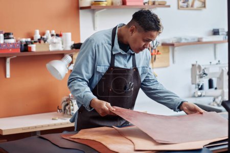 Téléchargez les photos : Portrait latéral du jeune artisan inspectant le cuir en atelier et créant des pièces faites à la main, espace de copie - en image libre de droit