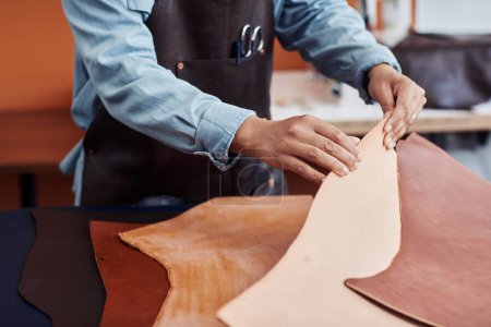 Téléchargez les photos : Gros plan d'un jeune artisan méconnaissable inspectant le cuir véritable en atelier et créant des pièces faites à la main, espace de copie - en image libre de droit