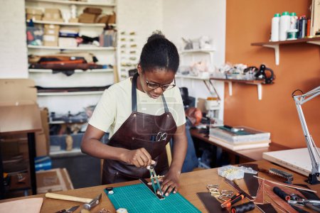 Téléchargez les photos : Portrait d'artisan femme noire mesurant une pièce en cuir dans un atelier de maroquinerie et créant des dessins faits à la main - en image libre de droit