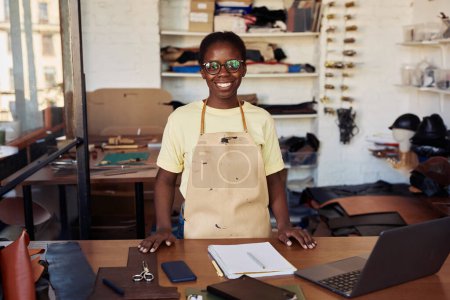 Portrait of female small business owner smiling at camera while posing in leatherworking workshop, copy space