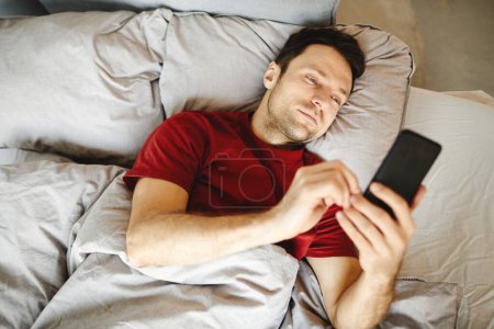 Photo for High angle view of mature man lying on bed in the morning and checking mail in his mobile phone - Royalty Free Image