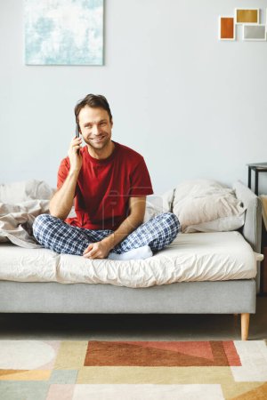 Téléchargez les photos : Jeune homme souriant assis sur son lit et parlant sur son téléphone portable le matin dans sa chambre - en image libre de droit