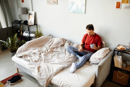Téléchargez les photos : Vue grand angle du jeune homme assis sur le lit et naviguant dans un ordinateur portable avec une tasse de café le matin - en image libre de droit