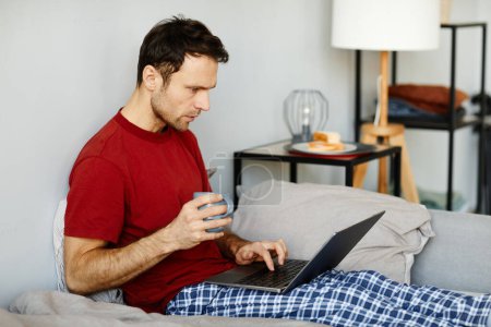 Foto de Joven sentado en la cama con taza de café y tener onlne comunicación en el ordenador portátil en su fin de semana en casa - Imagen libre de derechos