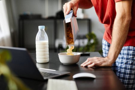 Téléchargez les photos : Gros plan du jeune homme qui met des flocons de maïs dans un bol pour les préparer avec du lait pour le petit déjeuner à table avec un ordinateur portable - en image libre de droit
