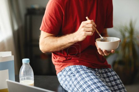 Téléchargez les photos : Gros plan du jeune homme tenant un bol et mangeant des céréales pour le petit déjeuner tout en regardant une vidéo sur ordinateur portable - en image libre de droit