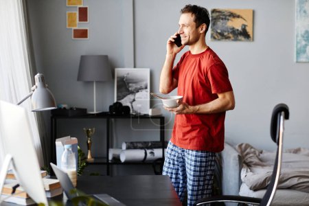 Photo for Young man in domestic clothes talking on mobile phone while having breakfast in the room - Royalty Free Image